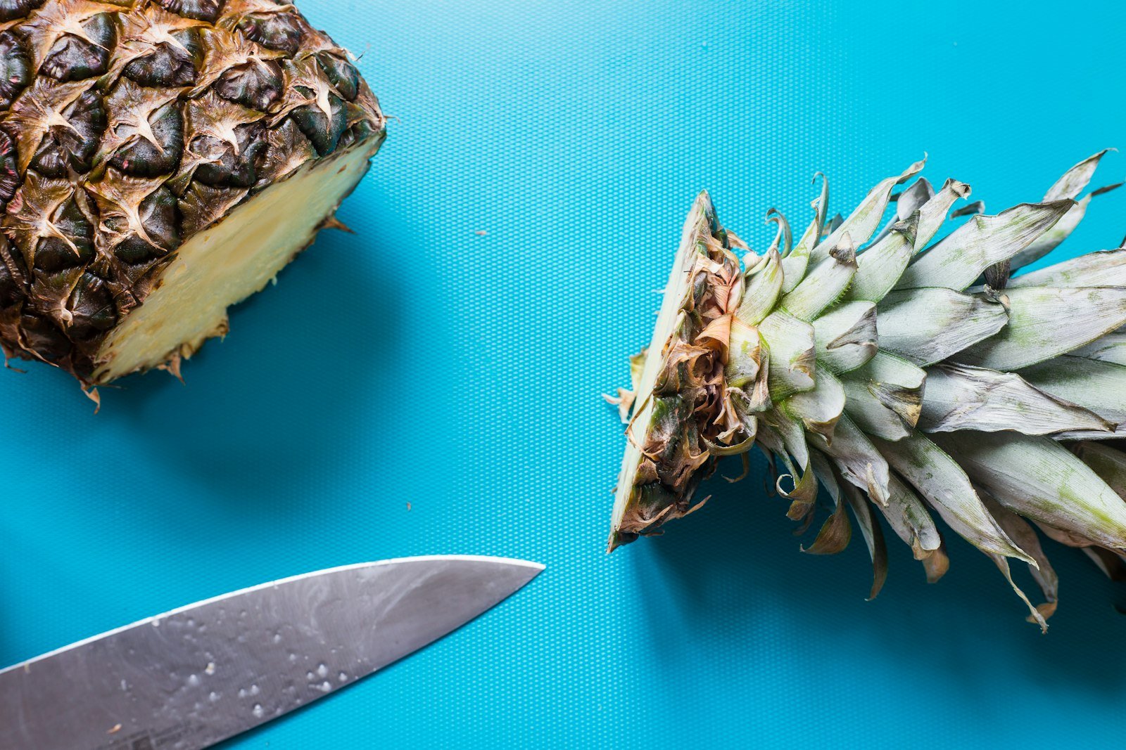 Abacaxi cortado ao meio com a coroa separada e faca de chef sobre a tábua de corte azul