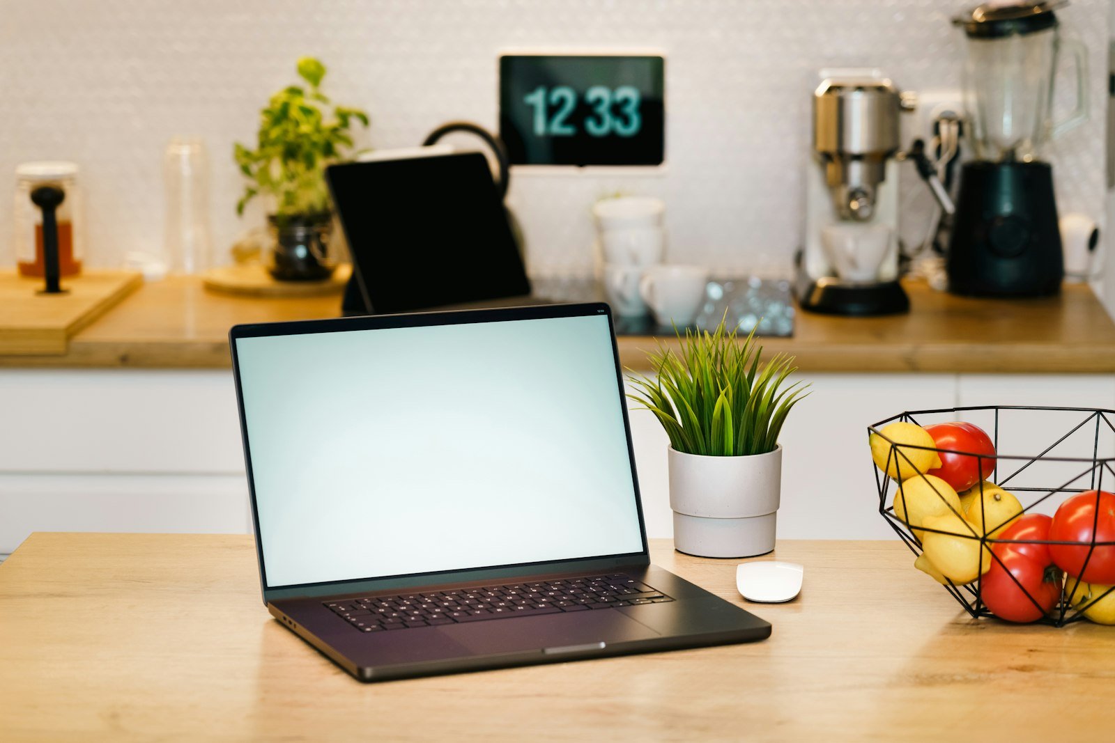 Notebook aberto sobre a bancada de madeira, com um tablet e a cafeteira ao fundo da cozinha